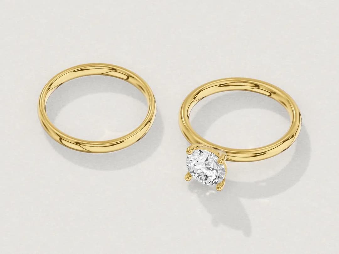 3D render of a yellow gold solitaire and a plain yellow gold band lying apart on a pale grey table.