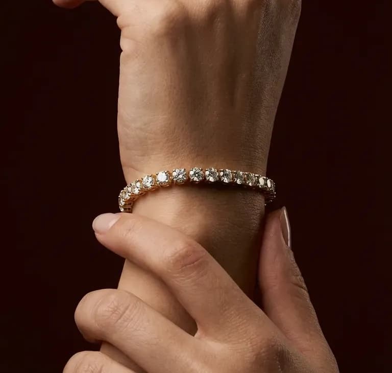 Photograph of a yellow gold diamond tennis bracelet on a wrist, held by the other hand against a dark background.