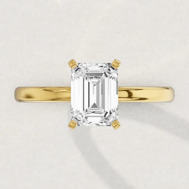 Face-up view of a yellow gold four-claw solitaire engagement ring on a 2 mm band with an emerald cut diamond the size of our 2 carat emerald cuts, about 8.6 by 6.0 mm, a 3D render drawn to the same scale as the other face-up rings on this page