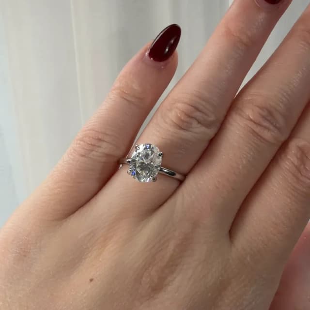 Photo of an engagement ring with a 2 carat oval and a hidden halo on a fine white metal band, worn on the ring finger of a hand with dark red nails against a white curtain