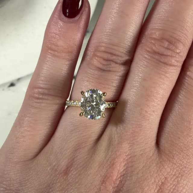 Photo of a yellow gold engagement ring with a 2 carat oval moissanite in four claws on a three-quarter pavé band, worn on the ring finger of a hand with dark red nails