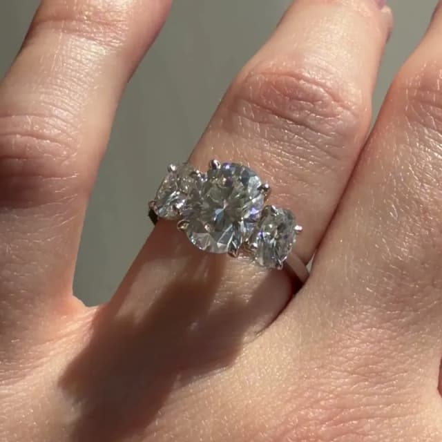 Close-up photo of the same platinum three-stone engagement ring with a 2 carat oval lab-grown diamond, worn on the ring finger in sunlight