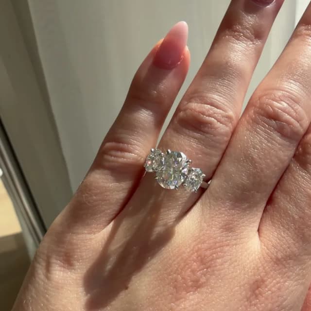 Photo of a platinum three-stone engagement ring with a 2 carat oval lab-grown diamond between two smaller side stones on a 2 mm band, worn on the ring finger in sunlight