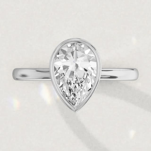 Face-up view of a white gold bezel solitaire engagement ring with a pear diamond the size of our 2 carat pears, a thin rim of metal around the whole stone including its point, a 3D render drawn to the same scale as the other face-up rings on this page